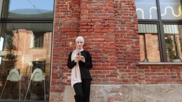 赤いレンガからの建物の壁の背景に立ってスマートフォンにヒジャーブのテキストで魅力的な笑顔のイスラム教徒の女性 市内の携帯電話を使用した若いアラビア人実業家の肖像画 — ストック動画