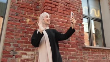Tesettürlü, tesettürlü, tesettürlü, akıllı telefonda kırmızı tuğladan duvar arkasında duran çekici bir müslüman kadın. Şehirde cep telefonu kullanan genç Arap iş kadınının portresi.