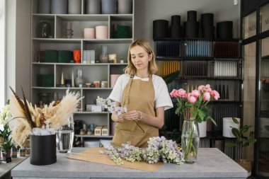 Portrait of young female florist holding violet flowers and looking at them. Professional and hardworking florist.