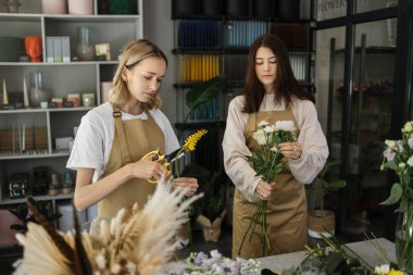 Beautiful caucasian florist girls making bouquet of flowers on table for sale against floral bokeh background in flower shop indoors. Two attractive females florists working in store.