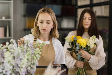 Two smiling female florists working behind counter in modern loft interior of floral shop. Floristics, business, decoration concept. Creative occupation in floral boutique.