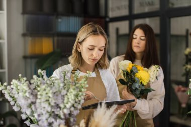 Creative female designer sit at desk in modern shop interior and make order for next period, woman assistant compose arrange plants flowers in bouquets and make floral compositions for sale.