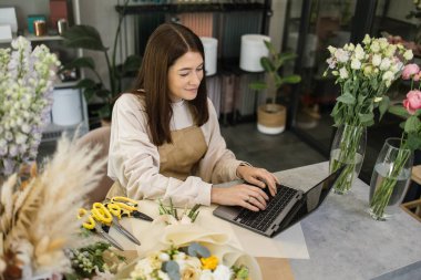 Portrait of attractive woman florist working on computer at workplace. Concentrated female sitting at counter in floral store and typing on laptop.