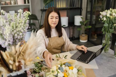 Portrait of attractive woman florist working on computer at workplace. Concentrated female sitting at counter in floral store and typing on laptop.