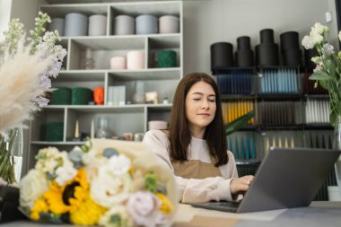 Çiçekçide dizüstü bilgisayarda çalışan önlüklü güzel kadın çiçekçinin portresi. Çekici bir kadın mağaza sahibi online siparişleri, e-postaları ve daktilo mesajlarını kontrol ediyor. E-ticaret, iş, internet kavramı