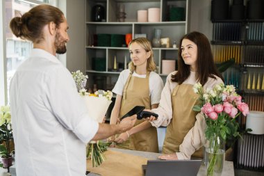 Çekici genç sakallı adam alışveriş yapıyor ve çiçekçide elektronik kredi kartıyla ödeme yapıyor. Erkek müşteri çiçek buketi için kartla ödeme yapıyor. Bahçe merkezinde alışveriş, iş..