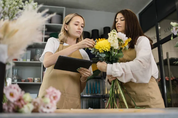 Team of two female florists in beige aprons using tablet while discussing which flowers to order for next week and making bouquet in modern interior floral shop.