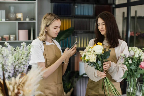 Two women florists making fashion modern bouquet of different flowers on counter and taking photo for social media. Masterclass. Gift for bride on wedding, mother, womens day.