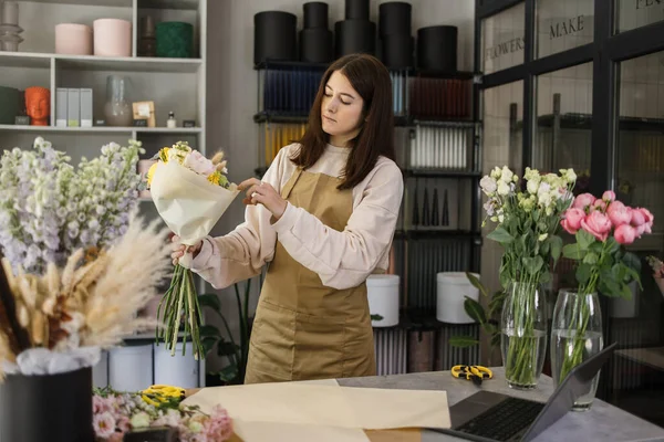 Portrait of young brunette female florist holding bouquet of flowers and looking at them. Professional and hardworking florist.