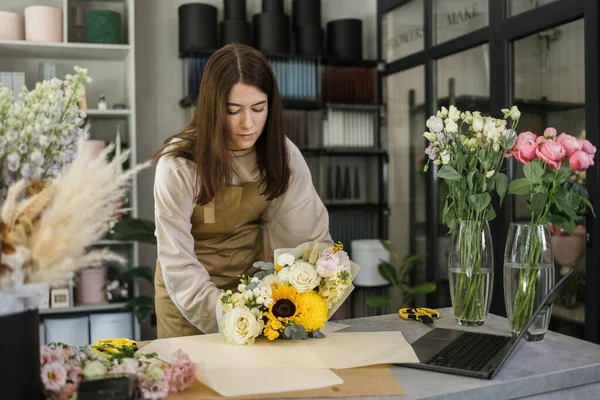 Portrait of young brunette female florist holding bouquet of flowers and looking at them. Professional and hardworking florist.