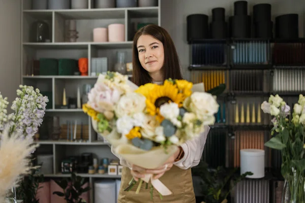 Smiling florist in uniform work in a flower shop behind counter with different varieties of flowers and huge bouquet in hands. Small business concept. Modern loft interior.