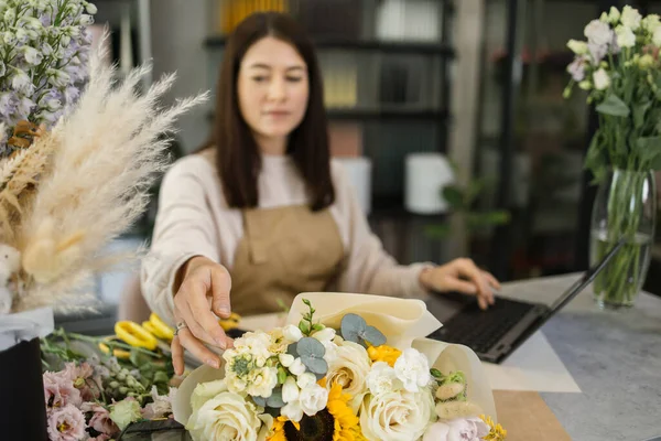 Beautiful flower shop owner wearing apron, working in store. Pretty young female florist using laptop to chat with client, customer, scroll news feed. Social media, technology, business concept.
