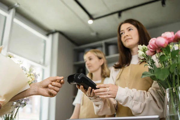 Two smiling woman florists holding card reader machine at counter with customer paying with credit card. Young florist shop assistant holding payment machine while buyer purchase a bunch flower.