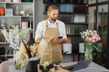 Küçük bir iş. Çiçekçide sakallı genç bir erkek çiçekçi akıllı telefon kullanarak müşteriye mesaj yazıyor. Çiçek tasarım stüdyosu, dekorasyon ve düzenlemeler. Çiçek teslimatı, yaratılış düzeni.