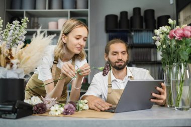 Two workers, man florist working on computer at workplace and woman helping him. Concentrated bearded male and contractor female sitting at counter in floral store and typing on laptop.