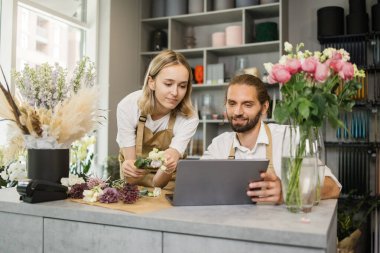 Two workers, man florist working on computer at workplace and woman helping him. Concentrated bearded male and contractor female sitting at counter in floral store and typing on laptop.