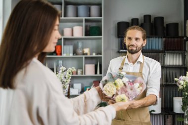 Attractive bearded man owner of flower shop advising female customer before purchase of flowers. Smiling florist giving bouquet to customer in flower shop.