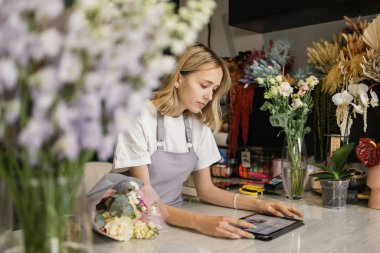 İş yerinde tablet üzerinde çalışan çekici kadın çiçekçinin portresi. Çiçekçide tezgahta oturmuş dijital tablette daktilo yazan konsantre bir kadın..