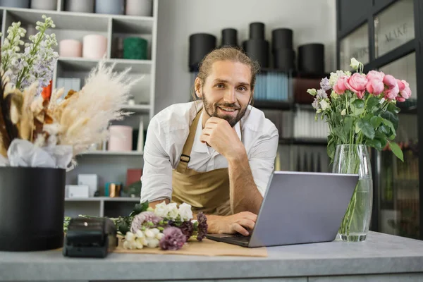Florist bearded man using laptop. Greenhouse worker, male botanist close-up, small business owner. Gardener working in flower shop, plant store.