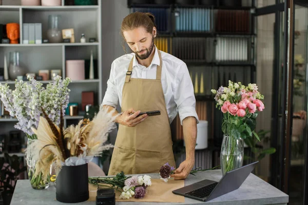 Portrait of young male florist making fashion modern bouquet of different flowers on counter and taking photo on smartphone for social media. Masterclass. Gift for bride on wedding, mother womens day