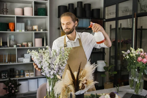 Smiling florist in uniform work in a flower shop behind counter taking care of different varieties of flowers by moisturizing them. Small business concept. Modern loft interior.