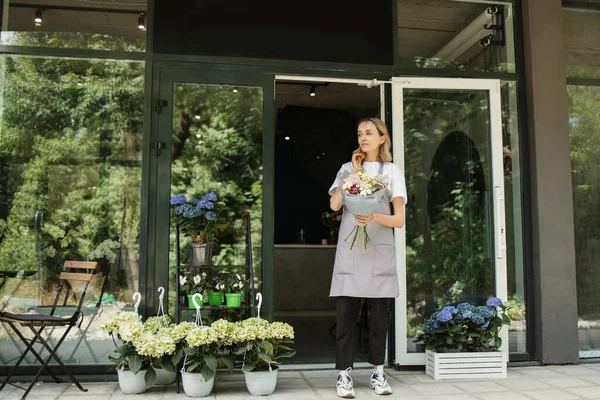 Business Woman near flower shop door. Photo of beautiful woman standing in front of flower shop. Female Florist Outside Shop. Woman working at flower shop smiling