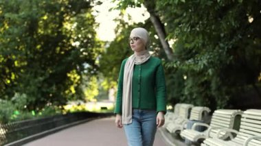 Smiling young muslim woman touching her eyeglass during break outdoor. Cheerful arabic woman in hijab walking in green summer city park.