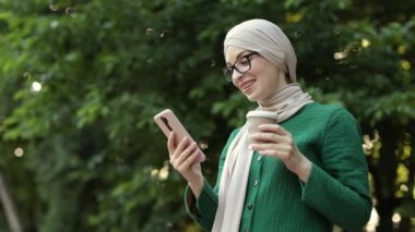 Neşeli genç İslamcı iş kadını şehir parkında yürürken cep telefonuyla konuşuyor ve kahve içiyor. Kahve molası sırasında akıllı telefon kullanan tesettürlü güzel genç bir kadın..