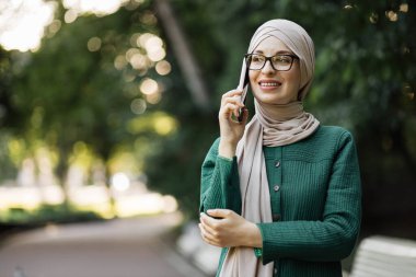 Gülümseyen müslüman bir kadının portresi. Telefon görüşmesi yapıyor. Mola sırasında ek iş alıyor. Parkta yürürken akıllı telefon kullanan tesettürlü neşeli Arap kadın..