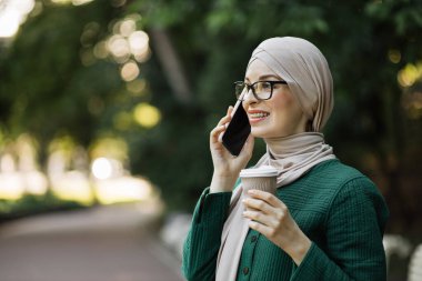 Neşeli genç İslamcı iş kadını şehir parkında yürürken cep telefonuyla konuşuyor ve kahve içiyor. Kahve molası sırasında akıllı telefon kullanan tesettürlü güzel genç bir kadın..