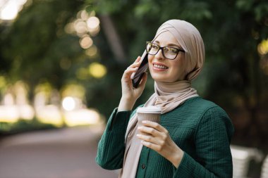 Neşeli genç İslamcı iş kadını şehir parkında yürürken cep telefonuyla konuşuyor ve kahve içiyor. Kahve molası sırasında akıllı telefon kullanan tesettürlü güzel genç bir kadın..