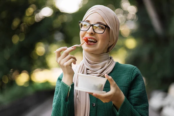 Portrait of a young attractive muslim woman eating salad at the background of park. Beautiful young islam female enjoying a fresh salad outdoor. Arab girl eating healthy meal.