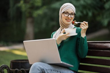 Keyifli Müslüman kız tesettürde sağlıklı salata yiyor parkta bankta oturuyor dizüstü bilgisayarda çalışıyor kahve molası sırasında dinleniyor, parkta yürüyor..