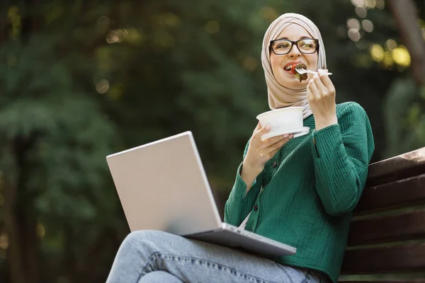 Happy smiling muslim businesswoman eating healthy salad on a break sitting on bench working on laptop. Female dieting nutrition concept. Attractive islam smiling girl enjoying veggie meal.