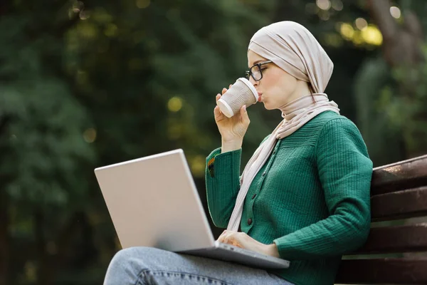 Happy smiling muslim business woman drinking coffee on a break sitting on bench working on laptop. Attractive islam smiling girl enjoying time during coffee break.