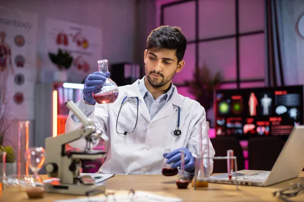 Yong indian male scientist working with test tubes wearing lab coat working in laboratory while examining biochemistry sample in test tube and scientific instruments.