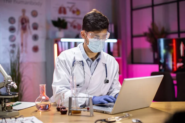 Biochemical research of scientist working with laptop for vaccine development in pharmaceutical laboratory. Indian man doctor observe virus evolution using high tech researching diagnosis.