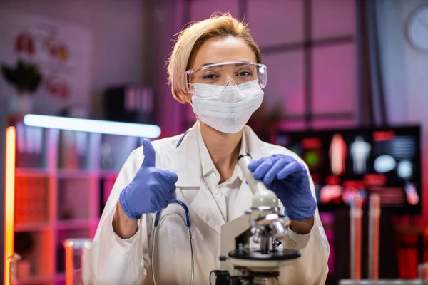 Experienced female scientist in health industry doing analysis of substance using microscope. Chemist researcher in sterile lab doing experiments for medical industry using modern technology, thumb up