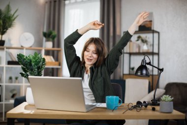 Excited caucasian businesswoman sitting at table with modern laptop and keeping arms raised for celebrating good news at work. Concept of success.
