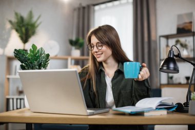 Attractive female freelancer with cup of coffee in casual outfit using modern laptop for work at home. Caucasian woman with dark hair sitting at table and typing on computer.