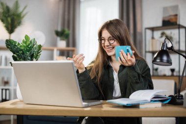 Excited young caucasian woman holding cup with hot drink and laptop for video conversation at office. Female freelancer sitting at table and taking notes during video chat.