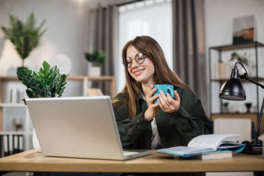 Happy laughing woman with cup of coffee talking and smiling during video chat on modern laptop. Pretty dark-haired female looking at funny movie using digital computer.