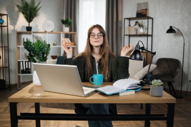 Pretty woman freelancer in eyeglasses sitting at table with closed eyes and relieving stress by meditation at workplace. Concept of relaxation and harmony, no stress free relief at work