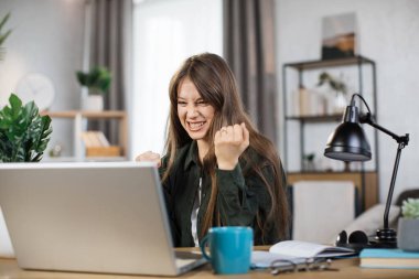 Happy caucasian woman student in casual wear sitting at table with modern laptop and keeping arms raised. Concept of people, technology and distance learning and success.