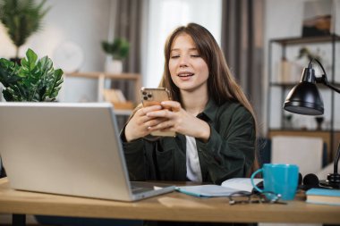 Confident young woman writing message during mobile conversation at office. Caucasian female worker with long dark hair sitting at table with opened laptop.