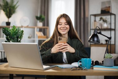 Confident young woman writing message during mobile conversation at office. Caucasian female worker with long dark hair sitting at table with opened laptop.