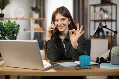 Young pretty woman in casual outfit talking on mobile phone and typing on laptop while sitting at workplace. Concept of corporate communication showing sign ok.