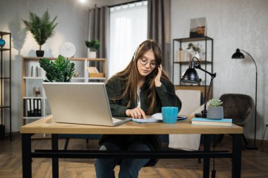 Young caucasian woman in headset sitting at desk with laptop and talking during video call. Businesswoman using modern devices for online conversation at work.