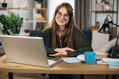 Young caucasian woman in headset sitting at desk with laptop and talking during video call. Businesswoman using modern devices for online conversation at work.