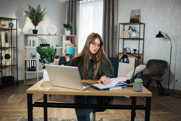 Smiling Caucasian woman in eyeglasses and denim shirt, posing indoors at bright office or home interior. Pretty lady with hot drink reading notes while working at laptop.